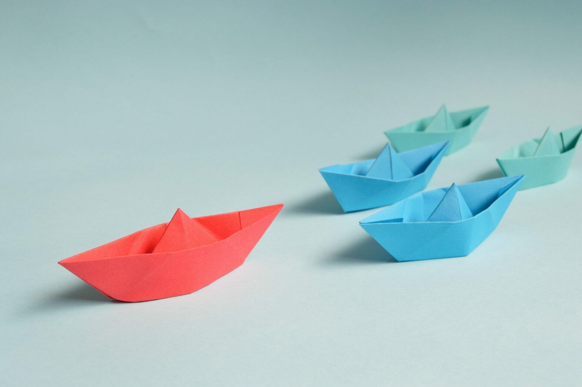 White backdrop, 3 blue paper boats are following one red paper boat